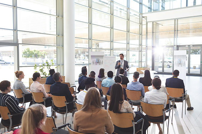 Egy csoport egy modern, világos konferenciateremben ül, és figyelmesen hallgatja az előadót; aki egy feliratozott whiteboard mellett áll.