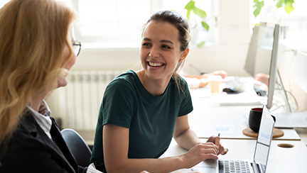 Zwei Personen sitzen an einem Schreibtisch in einem Büro; vor ihnen stehen ein Laptop, eine Kaffeetasse und Dokumente – eine Szene einer Arbeitsbesprechung.