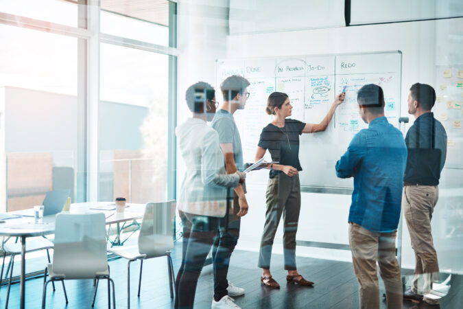 Munkatársak csoportja megbeszélést tart, a háttérben egy whiteboard segíti a közös munkát.