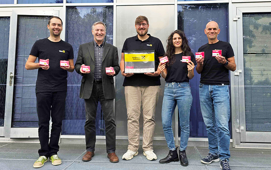 Öt személy látható egy épület előtt, Raspberry Pi dobozokat tartanak; egyikük egy Raiffeisen Software logóval ellátott kartondobozt tart maga előtt.