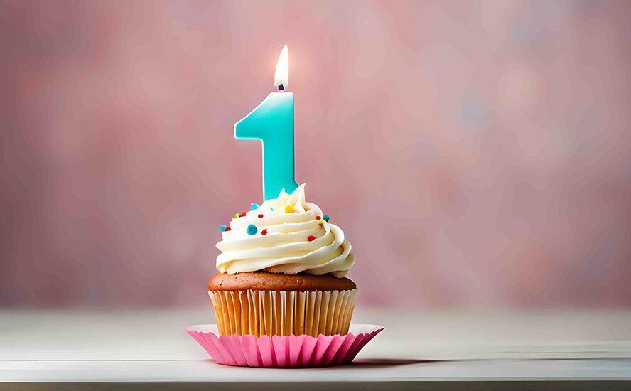 gy cupcake van a képen fehér krémmel és színes szórócukrokkal, tetején egy égő, egyes alakú gyertyával. A cupcake rózsaszín papírformában áll, elmosódott rózsaszín háttér előtt