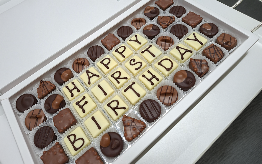 Nyitott bonbonos doboz; középen fehér csokoládékockákból formált betűk állnak, amelyek a „Happy First Birthday” feliratot adják ki. Körülötte különféle sötét és világos pralinék dekoratívan vannak elrendezve.